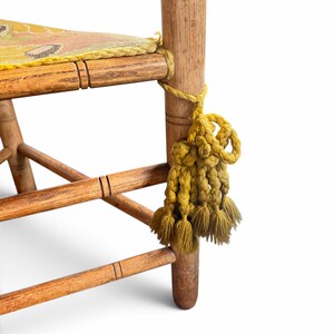 May include: A wooden chair featuring a seat upholstered in a yellow fabric with a multi-colored floral pattern. The chair frame is made of light brown wood with horizontal rungs and a decorative yellow tassel tied to the front leg.
