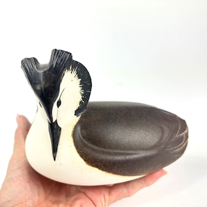 May include: A ceramic bird sculpture with a white body, a dark brown back, and a black crest. The bird's beak is black, and it is held in a person's hand. The sculpture is a decorative item.