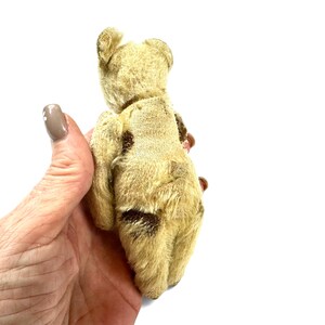 May include: A small, vintage-style teddy bear crafted from light beige, textured mohair fabric. The bear features visible patches of darker brown fur on its back and limbs, with rounded ears and a compact, articulated body structure.