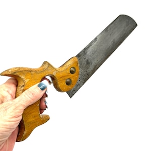 Könnte beinhalten: Eine Vintage-Handsäge mit einem verwitterten Holzgriff, einer ausgeprägten ergonomischen Form und zwei messingfarbenen Metallbefestigungen. Das Sägeblatt besteht aus dunklem, oxidiertem Stahl mit einer geraden, gezahnten Kante für Holzarbeiten.