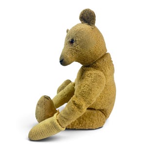 Pode incluir: Um urso de peluche de estilo vintage feito de tecido de pel&uacute;cia texturizado em tom castanho-dourado. O urso &eacute; apresentado numa posi&ccedil;&atilde;o sentada de perfil, com um nariz cosido em castanho escuro, pequenas orelhas redondas e olhos escuros brilhantes.
