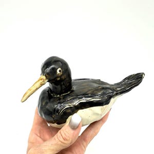 Op de afbeelding: Een keramieken vogelbeeldje met een zwarte kop en rug, een crème-kleurige snavel en een witte buik. De vogel is met de hand beschilderd en heeft een glanzende afwerking. Het beeldje is een decoratief item.