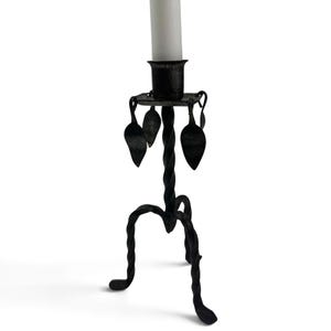 May include: A black metal candlestick holder featuring a twisted stem and three curved legs. The top platform includes a cylindrical candle cup and is decorated with four small, dangling metal leaf-shaped ornaments. A white candle is inserted.