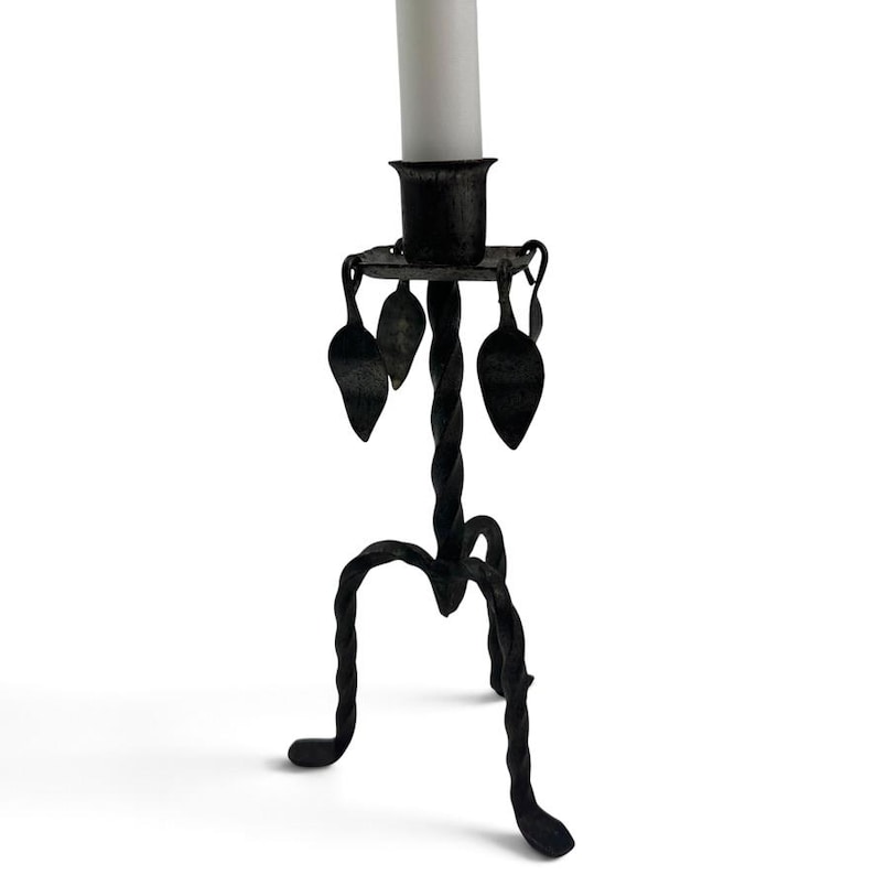 May include: A black metal candlestick holder featuring a twisted stem and three curved legs. The top platform includes a cylindrical candle cup and is decorated with four small, dangling metal leaf-shaped ornaments. A white candle is inserted.