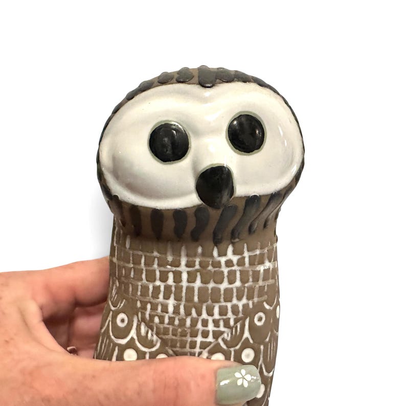 K&ouml;nnte beinhalten: Eine Eulenfigur aus Keramik mit einem wei&szlig;en Gesicht, gro&szlig;en schwarzen runden Augen und einem schwarzen Schnabel. Der K&ouml;rper ist braun mit einem texturierten Gittermuster auf der Brust und wei&szlig;en kreisf&ouml;rmigen Details an den Fl&uuml;geln. Dunkelbraune vertikale Streifen akzentuieren den Hals.
