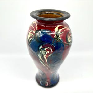 May include: A handcrafted ceramic vase with a glossy finish. The vase features a dark red base with blue and white swirling patterns. The rim is dark brown, and the interior is a lighter brown color. The vase has a bulbous shape and a narrow neck.