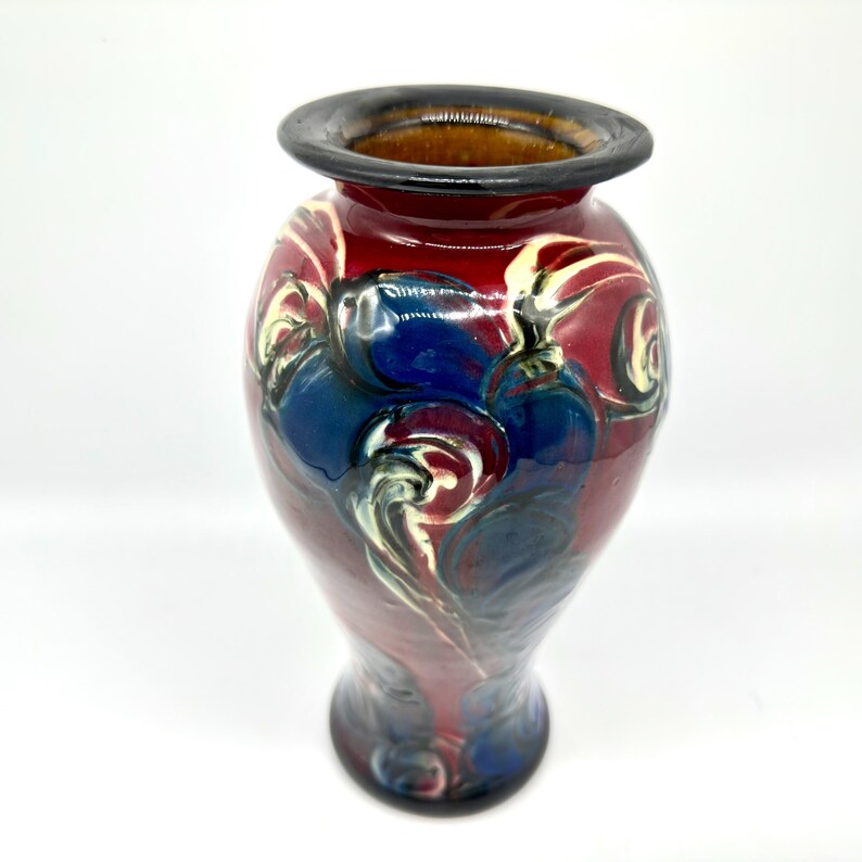 May include: A handcrafted ceramic vase with a glossy finish. The vase features a dark red base with blue and white swirling patterns. The rim is dark brown, and the interior is a lighter brown color. The vase has a bulbous shape and a narrow neck.