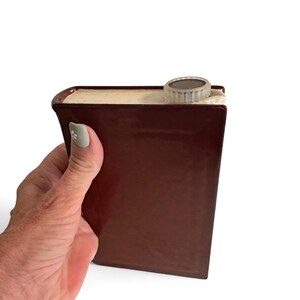 Puede incluir: Una petaca de cer&aacute;mica de color marr&oacute;n oscuro con forma de libro de tapa dura. La petaca presenta un acabado brillante, una zona del lomo texturizada que se asemeja a p&aacute;ginas de libro en color crema y un tap&oacute;n de rosca redondo de pl&aacute;stico.