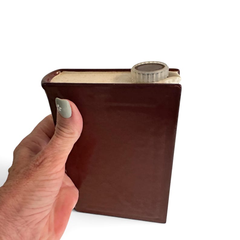 Puede incluir: Una petaca de cer&aacute;mica de color marr&oacute;n oscuro con forma de libro de tapa dura. La petaca presenta un acabado brillante, una zona del lomo texturizada que se asemeja a p&aacute;ginas de libro en color crema y un tap&oacute;n de rosca redondo de pl&aacute;stico.