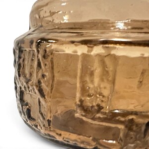 May include: A cylindrical glass container with a warm amber hue, featuring a thick, textured exterior surface with raised, irregular geometric patterns that create a rugged, tactile appearance across the translucent brown material.
