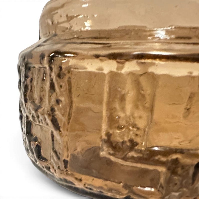 May include: A cylindrical glass container with a warm amber hue, featuring a thick, textured exterior surface with raised, irregular geometric patterns that create a rugged, tactile appearance across the translucent brown material.