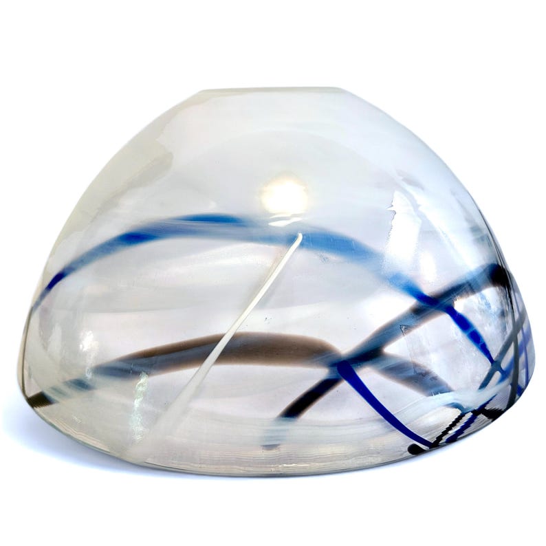 Pu&ograve; includere: Una ciotola in vetro trasparente dalla forma arrotondata e a cupola, caratterizzata da motivi lineari vorticosi blu e grigio scuro incorporati nel materiale. La ciotola presenta una superficie liscia e lucida con un'ampia apertura superiore.