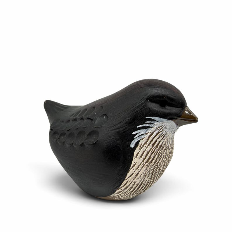 May include: A small, rounded bird figurine with a matte black body, featuring carved circular wing details and a textured, cream-colored chest with white accents. The bird has a small, pointed brown beak and a smooth, compact, stylized form.