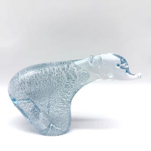 May include: A clear and light blue glass figurine shaped like a polar bear. The body features a textured, bubbled interior pattern, while the head and snout are smooth, transparent glass with a small blue dot for the eye.