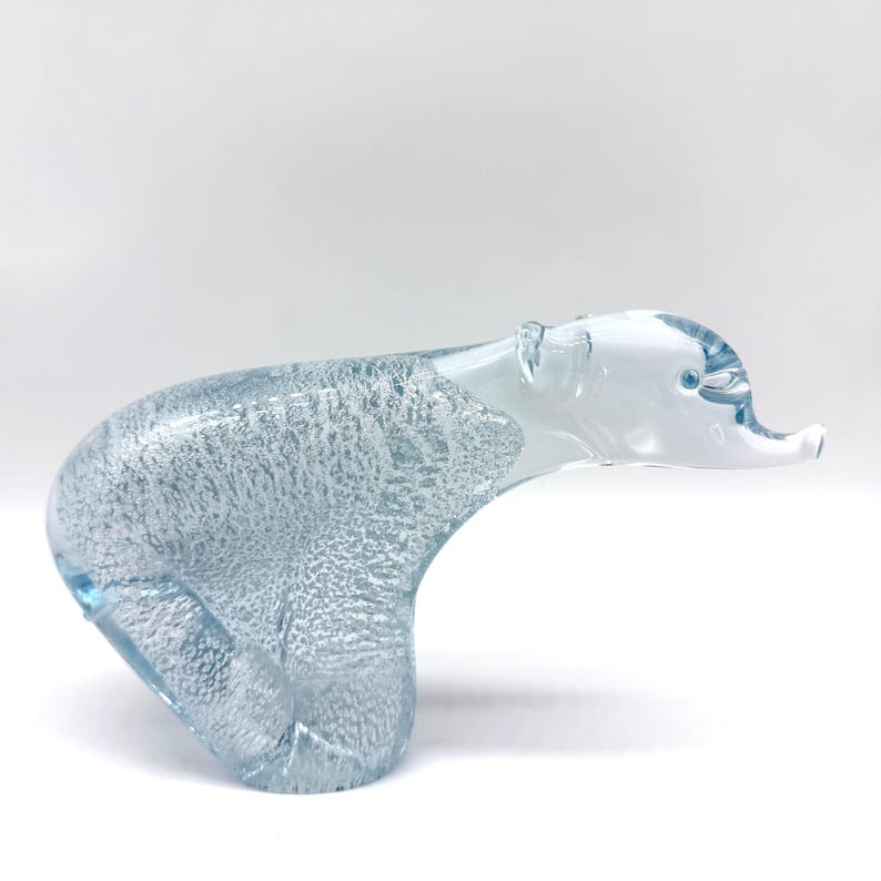May include: A clear and light blue glass figurine shaped like a polar bear. The body features a textured, bubbled interior pattern, while the head and snout are smooth, transparent glass with a small blue dot for the eye.