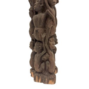 May include: A tall, dark brown wooden sculpture featuring a vertical stack of carved human figures. The figures are depicted in various poses, with some standing and others positioned above, creating a complex, textured, and layered cylindrical form.