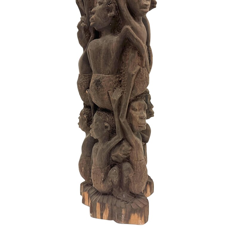 May include: A tall, dark brown wooden sculpture featuring a vertical stack of carved human figures. The figures are depicted in various poses, with some standing and others positioned above, creating a complex, textured, and layered cylindrical form.