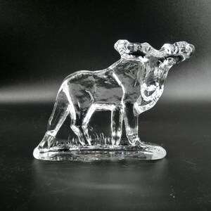 May include: A clear glass moose figurine. The moose is standing on a small base with a grassy texture. The antlers are detailed, and the glass is transparent, allowing light to pass through.