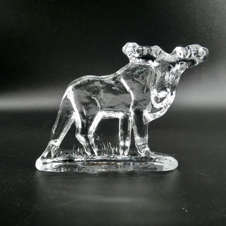 May include: A clear glass moose figurine. The moose is standing on a small base with a grassy texture. The antlers are detailed, and the glass is transparent, allowing light to pass through.