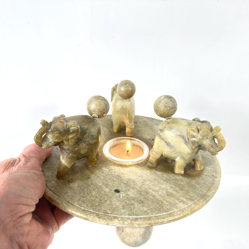 May include: A stone candle holder with three elephant figurines and three spherical accents. The candle holder has a circular base and a single central candle. The elephants are carved in detail and are a light beige color.