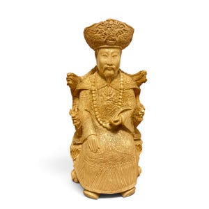 Op de afbeelding: Een lichtbruin gesneden beeldje van een zittende figuur met een versierde hoed en traditionele gewaden met ingewikkelde drakenmotieven. De figuur zit op een gedetailleerde stoel met gesneden armleuningen en draagt een ketting met kralen.