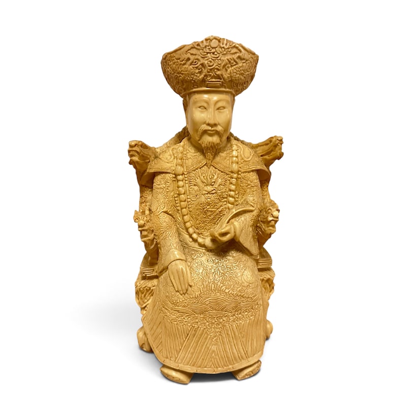 Op de afbeelding: Een lichtbruin gesneden beeldje van een zittende figuur met een versierde hoed en traditionele gewaden met ingewikkelde drakenmotieven. De figuur zit op een gedetailleerde stoel met gesneden armleuningen en draagt een ketting met kralen.