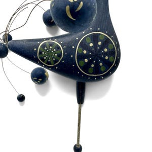 May include: A dark blue bird-shaped sculpture featuring hand-painted green and cream circular floral patterns on its body. The piece includes thin metal wires extending outward, each tipped with small dark blue spheres of varying sizes.