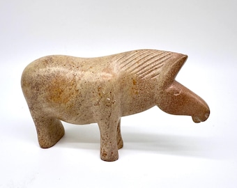 May include: A tan-colored, stylized horse sculpture. The sculpture has a rounded body, four legs, and a detailed head with carved lines representing the mane. The horse is standing on a white surface.