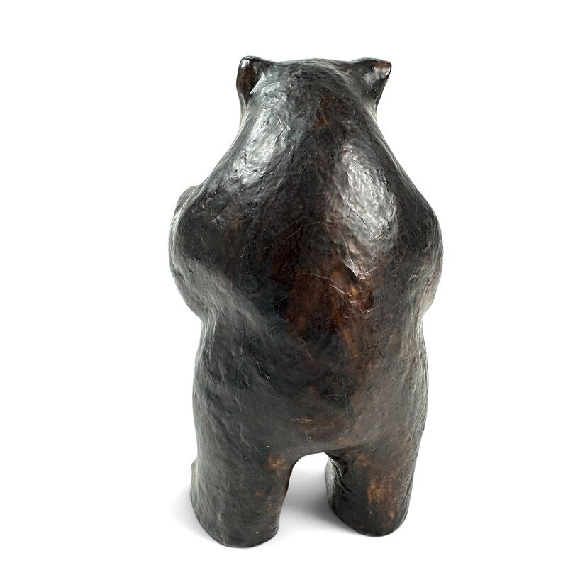 May include: A dark brown bear figurine shown from the back. The sculpture features a textured, uneven surface with deep brown and black tones, depicting the rounded shape of the bear's head, shoulders, and sturdy legs in a standing position.