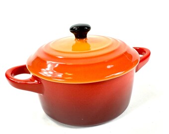 May include: A round enameled cast iron Dutch oven with a gradient finish transitioning from a deep red base to a bright orange lid. The pot features two side handles and a small, round black knob centered on the lid for lifting the cover.