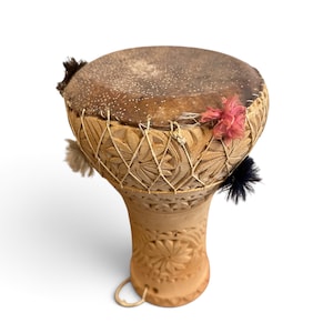 May include: A goblet-shaped drum with a natural skin head and a light brown clay body featuring intricate carved floral patterns. The drum is secured with a white cord lacing system and decorated with small, colourful fabric pom-poms in pink, black, and white.