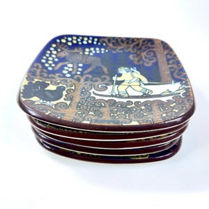 Op de afbeelding: Een stapel vierkante keramische borden met een donkerbruine rand. Elk bord heeft een blauwe achtergrond met een winters tafereel van een ski&euml;r, eland en vogel. De borden hebben een decoratieve, vintage stijl.