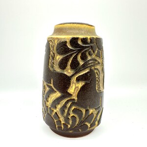 Peut inclure: Un vase en c&eacute;ramique marron avec une forme cylindrique et un dessus l&eacute;g&egrave;rement &eacute;vas&eacute;. Le vase pr&eacute;sente un motif en relief de chevaux et de feuillage dans une couleur marron plus claire. Le vase mesure environ 20 cm de haut.