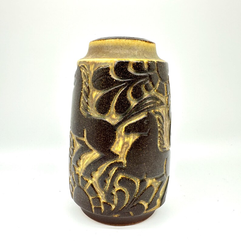 Peut inclure: Un vase en c&eacute;ramique marron avec une forme cylindrique et un dessus l&eacute;g&egrave;rement &eacute;vas&eacute;. Le vase pr&eacute;sente un motif en relief de chevaux et de feuillage dans une couleur marron plus claire. Le vase mesure environ 20 cm de haut.