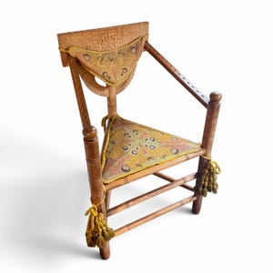 May include: A wooden chair with carved geometric patterns on the backrest, featuring a triangular seat and a small back cushion upholstered in a tan and yellow moth-patterned fabric, accented with yellow braided trim and hanging tassels.