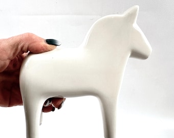 May include: A cream-colored ceramic horse figurine. The horse has a smooth, glossy finish and a minimalist design. The figurine is a stylized representation of a horse, with a simple body, legs, and head. The horse is approximately 6 inches tall.