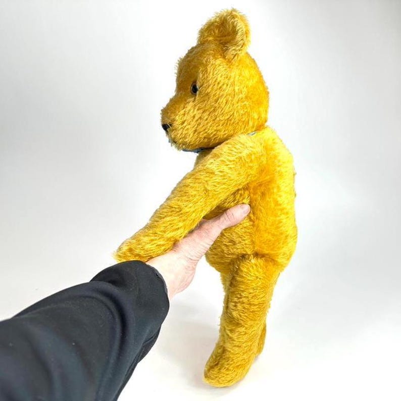 May include: A vintage golden yellow teddy bear with a classic design. The plush toy has a soft, textured appearance and is held by a person. The bear's features include a small black nose and button eyes. A nostalgic collectible.