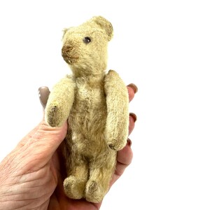 May include: A small, vintage-style teddy bear crafted from light beige, textured fur. The bear features a stitched brown nose, dark bead eyes, and articulated limbs. Its fur appears worn and aged, with a compact, upright posture and rounded ears.
