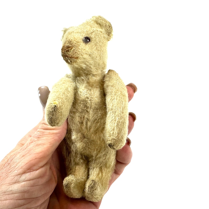 May include: A small, vintage-style teddy bear crafted from light beige, textured fur. The bear features a stitched brown nose, dark bead eyes, and articulated limbs. Its fur appears worn and aged, with a compact, upright posture and rounded ears.