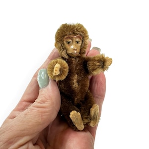 May include: A miniature monkey figurine crafted from brown fuzzy pipe cleaner material. The monkey features a molded plastic face with painted facial details, including eyes, a nose, and a mouth. It has small, light-colored felt hands and feet.