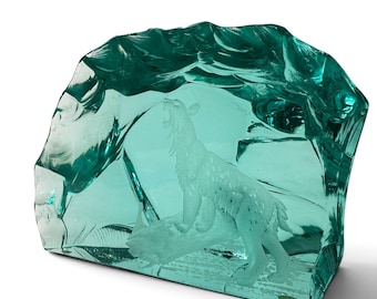 May include: A translucent teal glass sculpture featuring a carved lynx standing on a log. The glass has a thick, irregular, and textured outer edge that mimics the appearance of a natural ice formation, encasing the detailed feline figure.