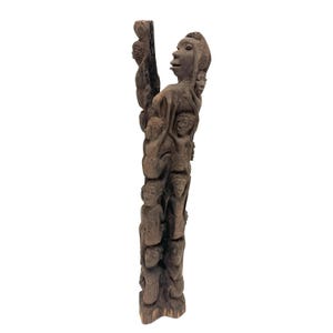 May include: A tall, dark brown wooden sculpture carved from a single piece of wood, featuring a vertical arrangement of multiple human figures stacked and intertwined, with a larger figure at the top and smaller figures positioned below.