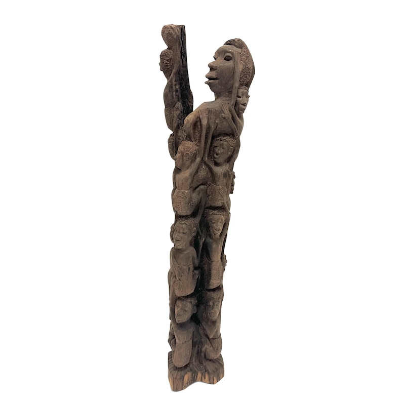 May include: A tall, dark brown wooden sculpture carved from a single piece of wood, featuring a vertical arrangement of multiple human figures stacked and intertwined, with a larger figure at the top and smaller figures positioned below.
