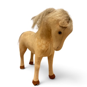 May include: A tan-colored plush horse toy with a soft, felt-like texture. The horse features a light-colored mane and tail made of synthetic hair, dark brown hooves, and small, dark, round eyes. The body is constructed with visible seams.