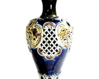 Puede incluir: Un jarrón de cerámica alto con un cuerpo esmaltado en azul profundo y adornos en relieve de color crema y marrón. El diseño incluye un patrón de celosía central, volutas ornamentales y un borde acampanado marrón oscuro.