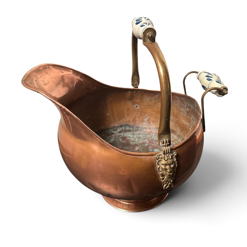 Pu&ograve; includere: Un secchio per carbone in rame con finitura lucida, dotato di un manico in ottone con impugnatura in ceramica bianca decorata con motivi floreali blu. Il manico &egrave; fissato al corpo con supporti in ottone a forma di testa di leone.