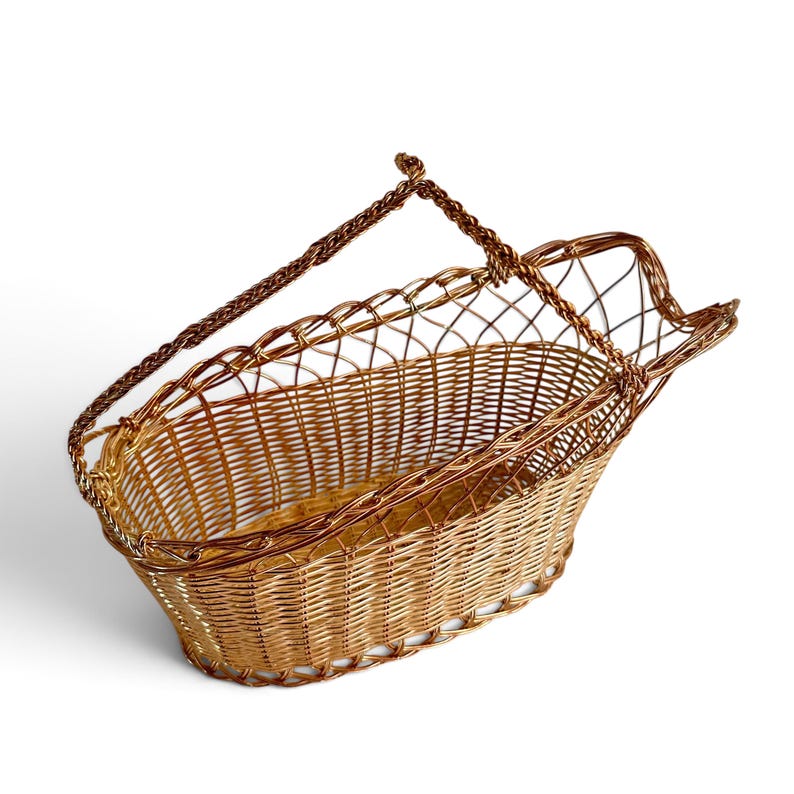 Puede incluir: Una cesta de alambre de color cobre con base ovalada y un dise&ntilde;o de rejilla abierta. La cesta cuenta con una sola asa de alambre trenzado unida a los lados, creando un recipiente estructurado y ligero con un acabado pulido.