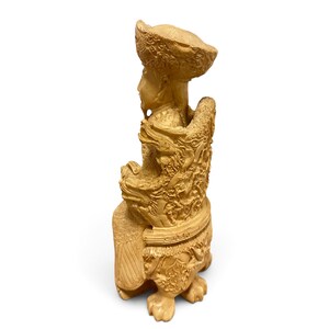 Op de afbeelding: Een lichtbeige uitgesneden beeldje van een zittende figuur met een hoed met brede rand en versierde gewaden. De figuur zit op een gedetailleerde stoel met drakenmotieven en patronen, rustend op uitgesneden poten met klauwen.