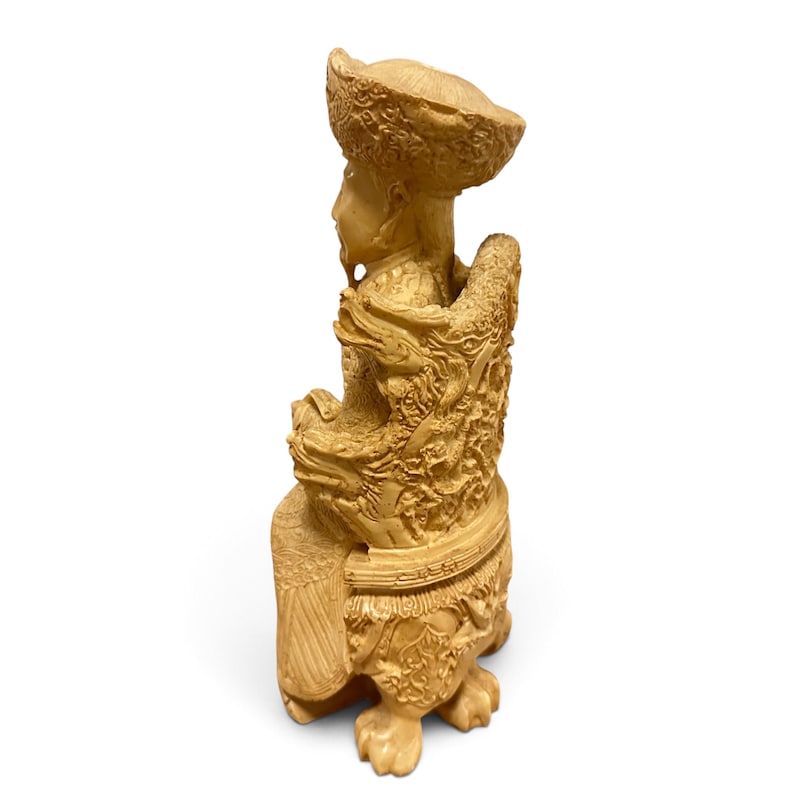 Op de afbeelding: Een lichtbeige uitgesneden beeldje van een zittende figuur met een hoed met brede rand en versierde gewaden. De figuur zit op een gedetailleerde stoel met drakenmotieven en patronen, rustend op uitgesneden poten met klauwen.