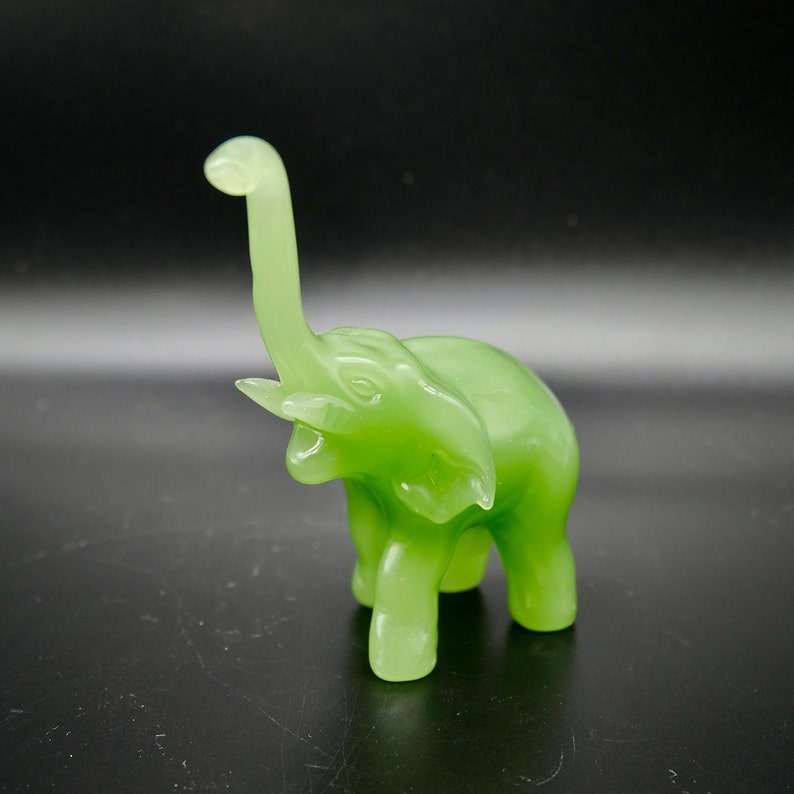 K&ouml;nnte beinhalten: Eine durchscheinende, hellgr&uuml;ne Elefantenfigur mit erhobenem R&uuml;ssel. Der Elefant hat detaillierte Merkmale, einschlie&szlig;lich Ohren und Sto&szlig;z&auml;hne, und steht auf vier Beinen. Die Figur besteht aus einem glatten, gl&auml;nzenden Material.